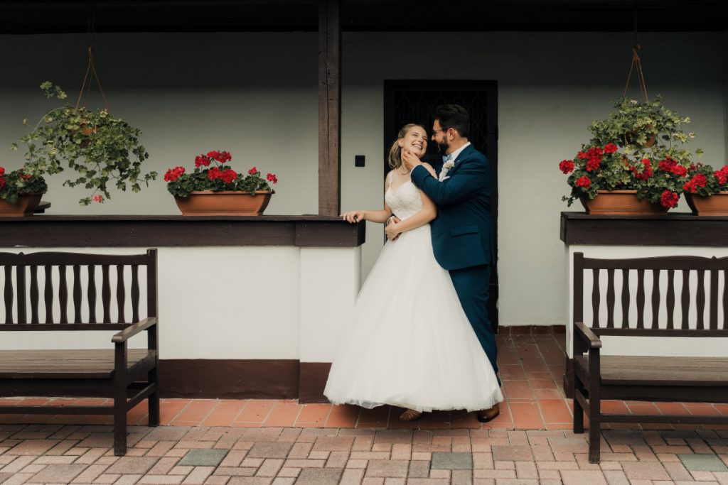 Kreatív esküvői fotózás Biczó Csárdában, fiatal pár muskátlik között, romantikus pillanatok a korabeli épület előtt